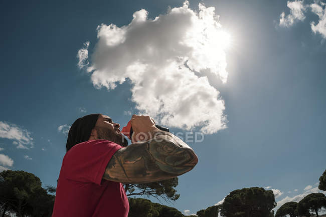Человек пьет во время тренировки против голубого неба с облаками — стоковое фото