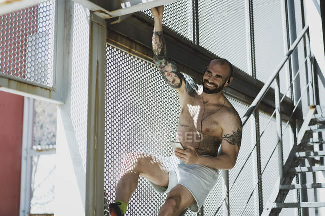 Handsome shirtless athlete smiling and listening to music from smartphone while hanging on bar near staircase during outdoor workout — Stock Photo