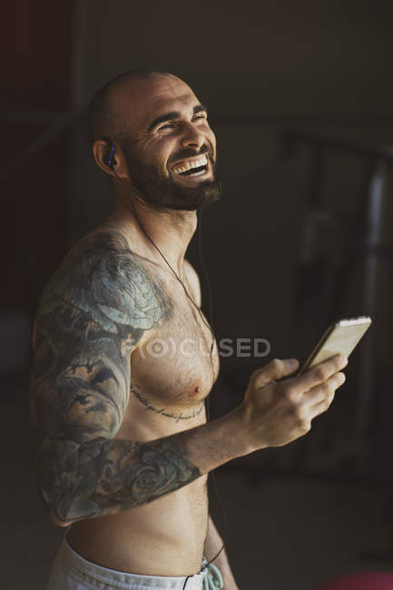 Hombre guapo tatuado con teléfono inteligente riendo y escuchando música mientras está de pie en el gimnasio moderno durante el entrenamiento - foto de stock
