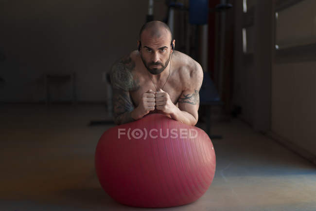 Guapo barbudo mirando a la cámara mientras realiza ejercicio de tablón en pelota suiza en el gimnasio - foto de stock