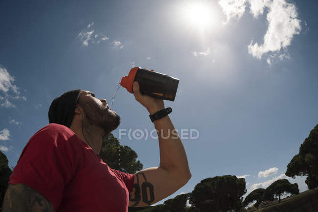 Homme buvant pendant l'entraînement contre le ciel bleu avec des nuages — Photo de stock