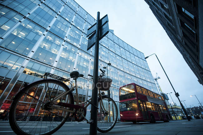 Bicycle parked near signpost against modern building and double-decker red bus on illuminated street of London — Stock Photo