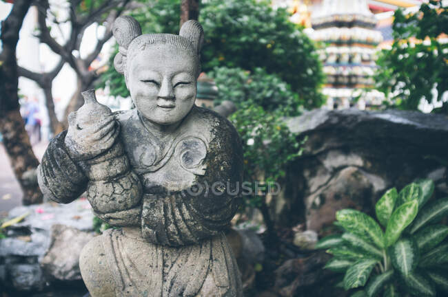 Прекрасная выветриваемая статуэтка, размещенная возле скал и зеленых тропических растений в удивительном саду в солнечный день в Таиланде — стоковое фото