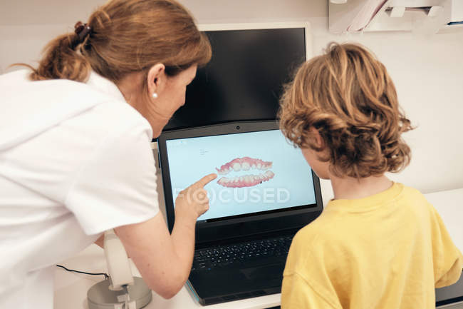 Female dentist demonstrating teeth scan on laptop screen to little boy in modern clinic — Stock Photo
