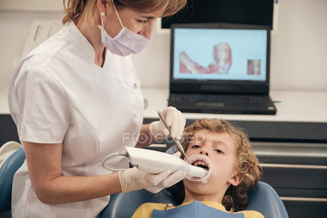 Frau in Maske und Arztuniform macht Scan der Zähne eines kleinen Jungen während der Arbeit in der Zahnarztpraxis — Stockfoto