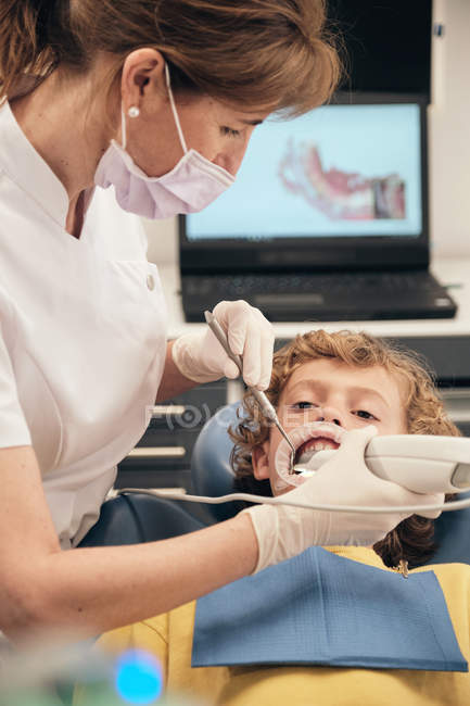 Mujer con máscara y uniforme médico haciendo escaneo de dientes de niño pequeño mientras trabajaba en la clínica dentista - foto de stock