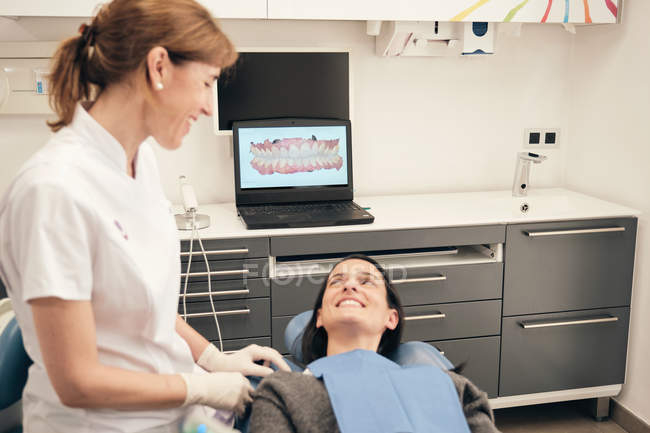 Dentista professionista in uniforme che parla con una donna sorridente mentre lavora in una clinica moderna — Foto stock