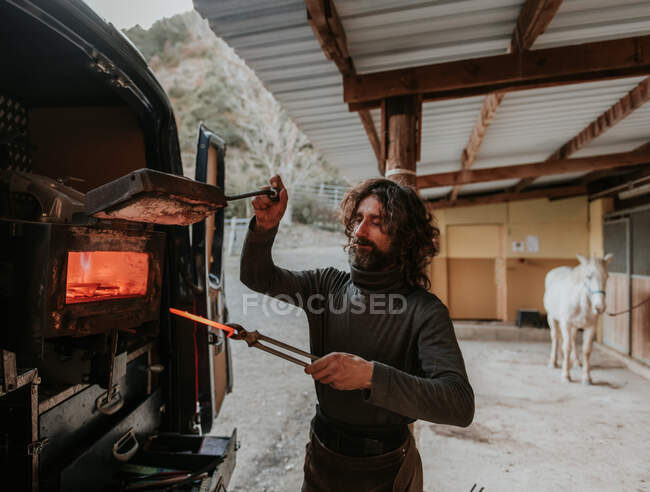 Bearded adult farrier taking hot horseshoe from portable furnace in back of car while working near stable on ranch — Stock Photo