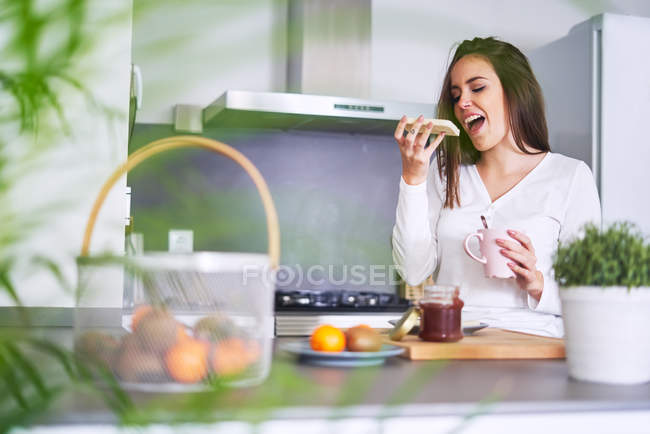 Junge glückliche Frau isst zu Hause Sandwich am Tisch in der Küche — Stockfoto