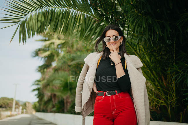 Smiling young woman in trendy outfit touching lips and smiling while standing near tropical palm leaves on street — Stock Photo