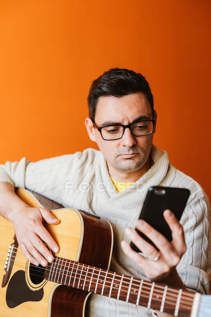 Людина з гітарою використовує мобільний телефон проти помаранчевої стіни — стокове фото