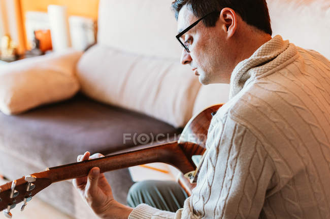 Крупный план человека в пуловере и очках, играющего дома на гитаре — стоковое фото