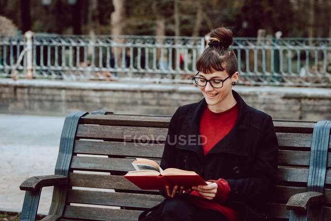 Young smiling elegant woman in eyeglasses reading book and sitting on bench in city park — Stock Photo