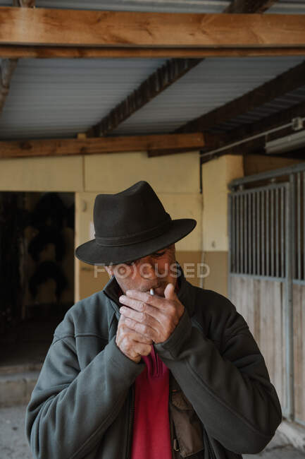 Uomo adulto in cappello fumare sigaretta mentre in piedi vicino bancarelle all'interno stalla su ranch — Foto stock
