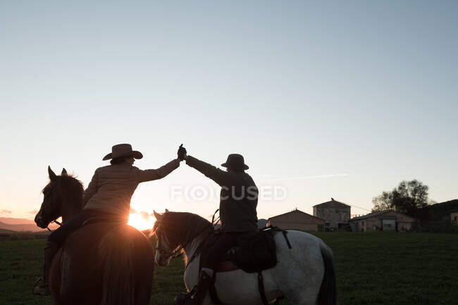 Vista trasera del hombre y la mujer montar a caballo y dar cinco de altura entre sí contra el cielo puesta del sol en el rancho - foto de stock