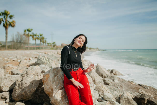 Mulher com olhos fechados em mar calmo apoiando-se na rocha na praia ensolarada — Fotografia de Stock