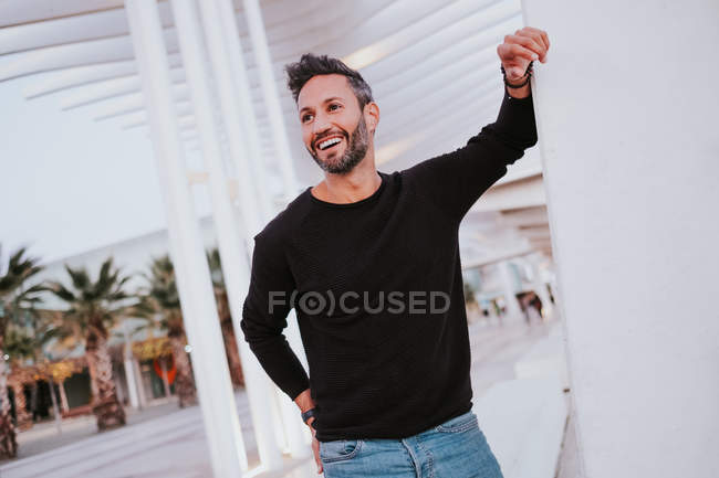 Adult handsome elegant cheerful male in casual wear leaning on grey wall and looking away on city street — Stock Photo