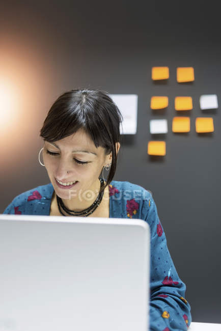 Lächelnde Geschäftsfrau arbeitet am Laptop, während sie im modernen Büro am Schreibtisch sitzt — Stockfoto