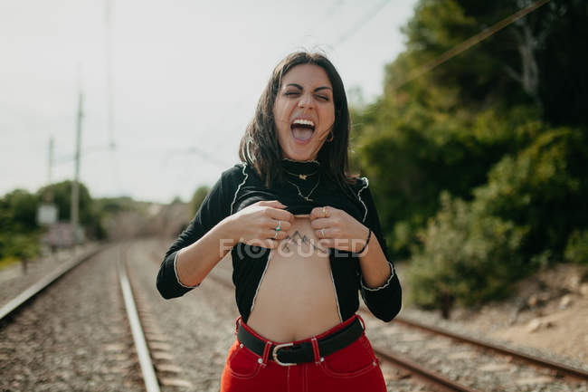 Young woman in stylish outfit screaming and lifting sweater to show mountains tattoo while standing on blurred background of rails — Stock Photo