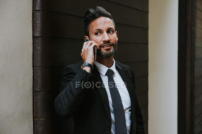 Adulto bell'uomo d'affari elegante in abito formale guardando altrove e parlando sul telefono cellulare vicino alla parete — Foto stock