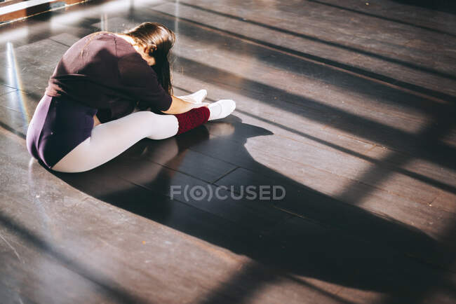 Visão traseira da mulher sentada no chão ensolarado do estúdio e aquecendo o corpo antes da dança. — Fotografia de Stock