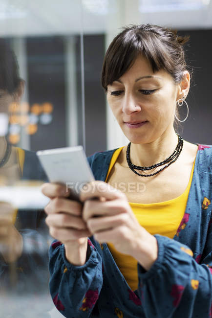Managerin nutzt Smartphone, während sie an Glaswand in modernem Büro steht — Stockfoto