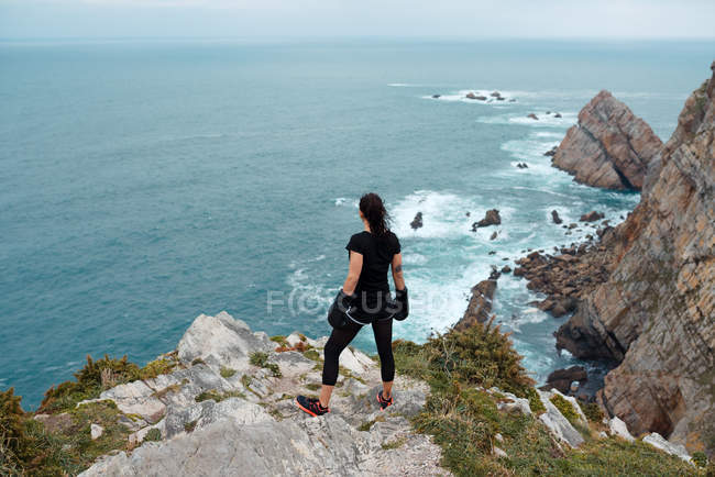 Back view of woman in boxing gloves standing on stony cliff and looking at sea during outdoor training — Stock Photo