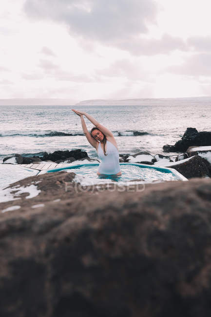 Молода жінка з закритими очима відпочиває у воді басейну біля скель і хмарного неба на морському узбережжі — стокове фото