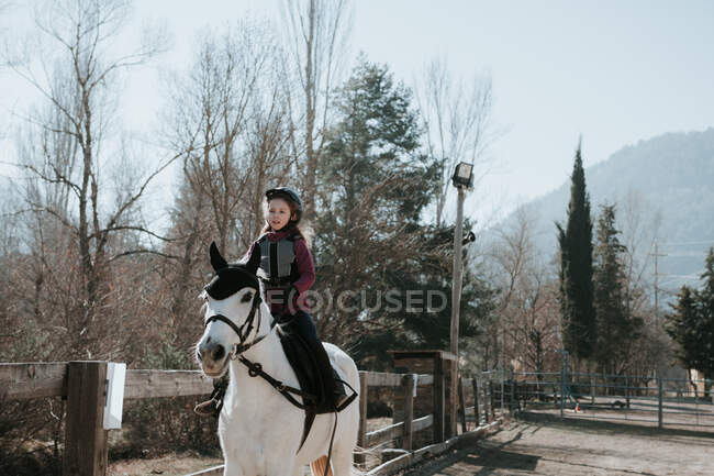 Doce menina no capacete equitação obediente cavalo branco no recinto durante a lição no dia de outono na fazenda — Fotografia de Stock