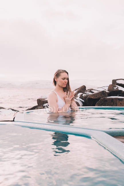 Giovane donna con gli occhi chiusi meditando in acqua di piscina vicino rocce e cielo nuvoloso — Foto stock