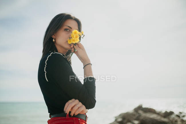 Чуттєва молода жінка з купою жовтих квітів, що стоять біля моря в сонячний день — стокове фото