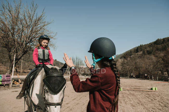 Молодая женщина в шлеме учит очаровательную маленькую девочку ездить на лошади во время урока в солнечный осенний день на ранчо — стоковое фото