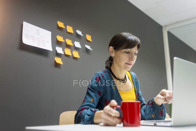 Femme d'affaires avec tasse de boisson chaude navigation ordinateur portable tout en travaillant dans le bureau moderne — Photo de stock