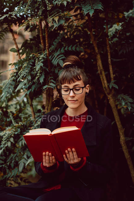 Joven atractiva mujer elegante en anteojos libro de lectura y sentado en el banco en el jardín de la ciudad - foto de stock