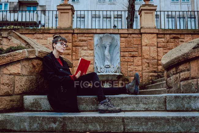 Jovem mulher elegante em óculos leitura livro e sentado em escadas na frente do edifício na cidade — Fotografia de Stock