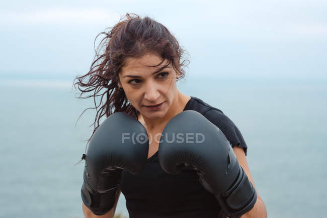 Mujer adulta en ropa deportiva practicando golpes durante el entrenamiento de kickboxing cerca del mar - foto de stock
