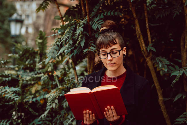 Young elegant woman in eyeglasses reading book in city garden — Stock Photo