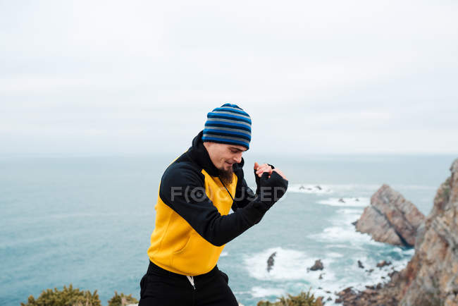 Adult bearded man in sportswear practicing punches during kickboxing workout on rocky cliff near sea — Stock Photo