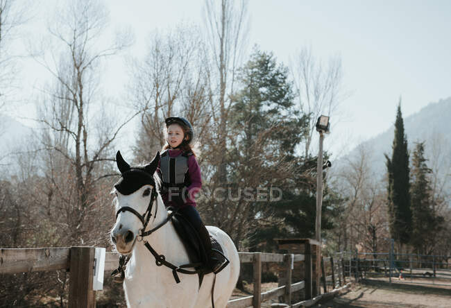 Süßes kleines Mädchen im Helm reitet gehorsam weißes Pferd im Gehege während des Unterrichts am Herbsttag auf der Ranch — Stockfoto