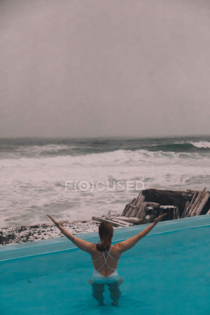 Vista posteriore di giovane donna con mani alzate che riposano in acqua di piscina vicino a scogliere sulla costa e mare tempestoso — Foto stock