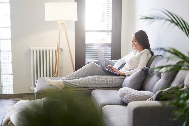 Giovane donna sorridente che utilizza il computer portatile e si riposa sul divano a casa — Foto stock