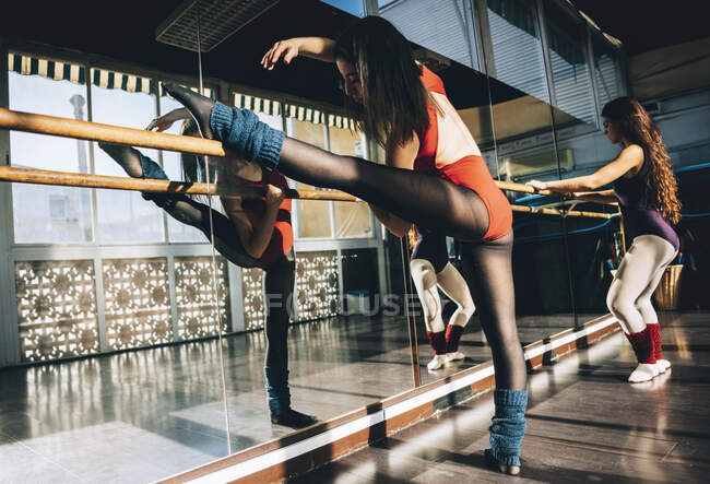 Confident young sportive women in bodysuits practicing in ballet studio stretching with barre. — Stock Photo