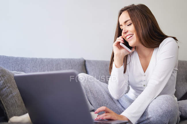 Smiling young happy woman using laptop and talking on mobile phone on sofa at home — Stock Photo