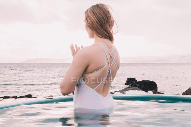 Молодая женщина медитирует в воде бассейна возле скал и облачного неба — стоковое фото