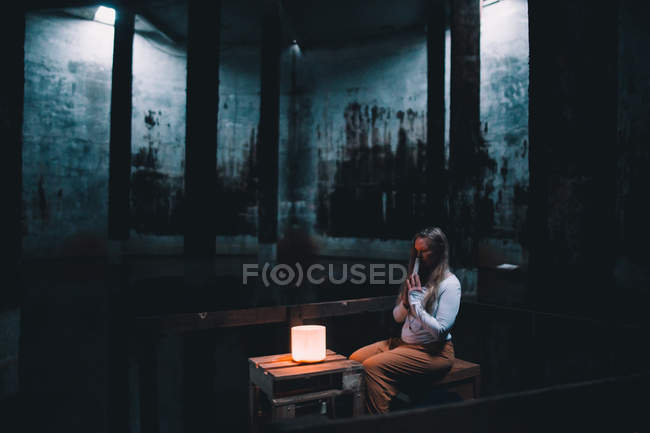 Молода жінка з молитовними руками, що сидить біля вогнів у темній будівлі — стокове фото