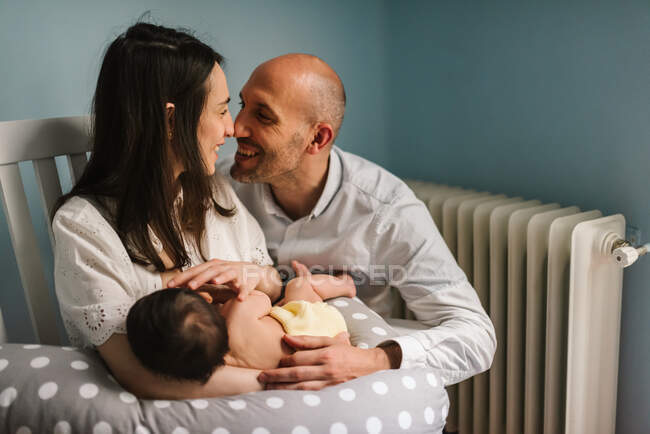 Homem adulto alegre abraçando mulher sorridente e bebê durante a amamentação em um berçário acolhedor em casa — Fotografia de Stock