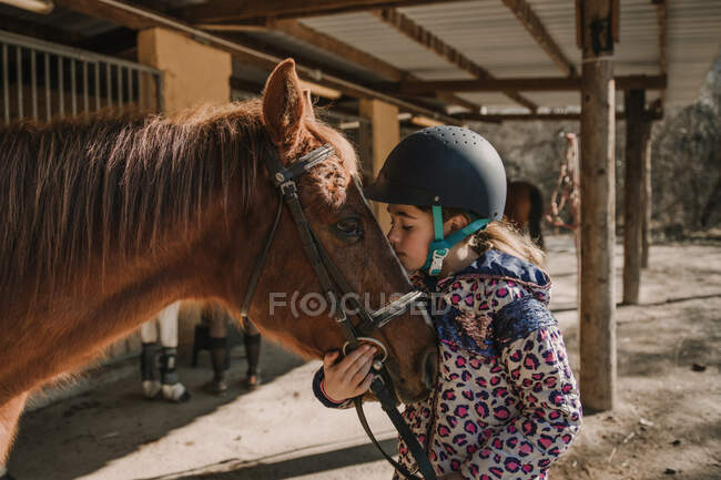 Carino bambina in casco baciare un cavallo bianco mentre in piedi vicino bancarelle in stalla durante la lezione di equitazione sul ranch — Foto stock