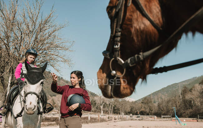Young female in helmet teaching adorable little girl to ride horse during lesson on sunny autumn day on ranch — Stock Photo