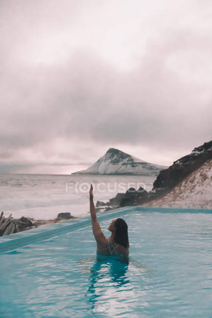 Вид на молодую женщину с поднятой рукой, отдыхающую в воде бассейна возле скал на побережье и бурное море — стоковое фото