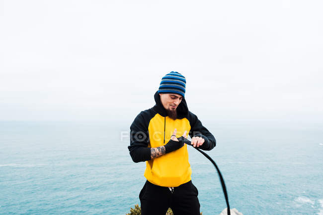 Бородатий дорослий чоловік обмотує пов'язку навколо руки, стоячи проти моря під час тренування з кікбоксингу на відкритому повітрі — стокове фото